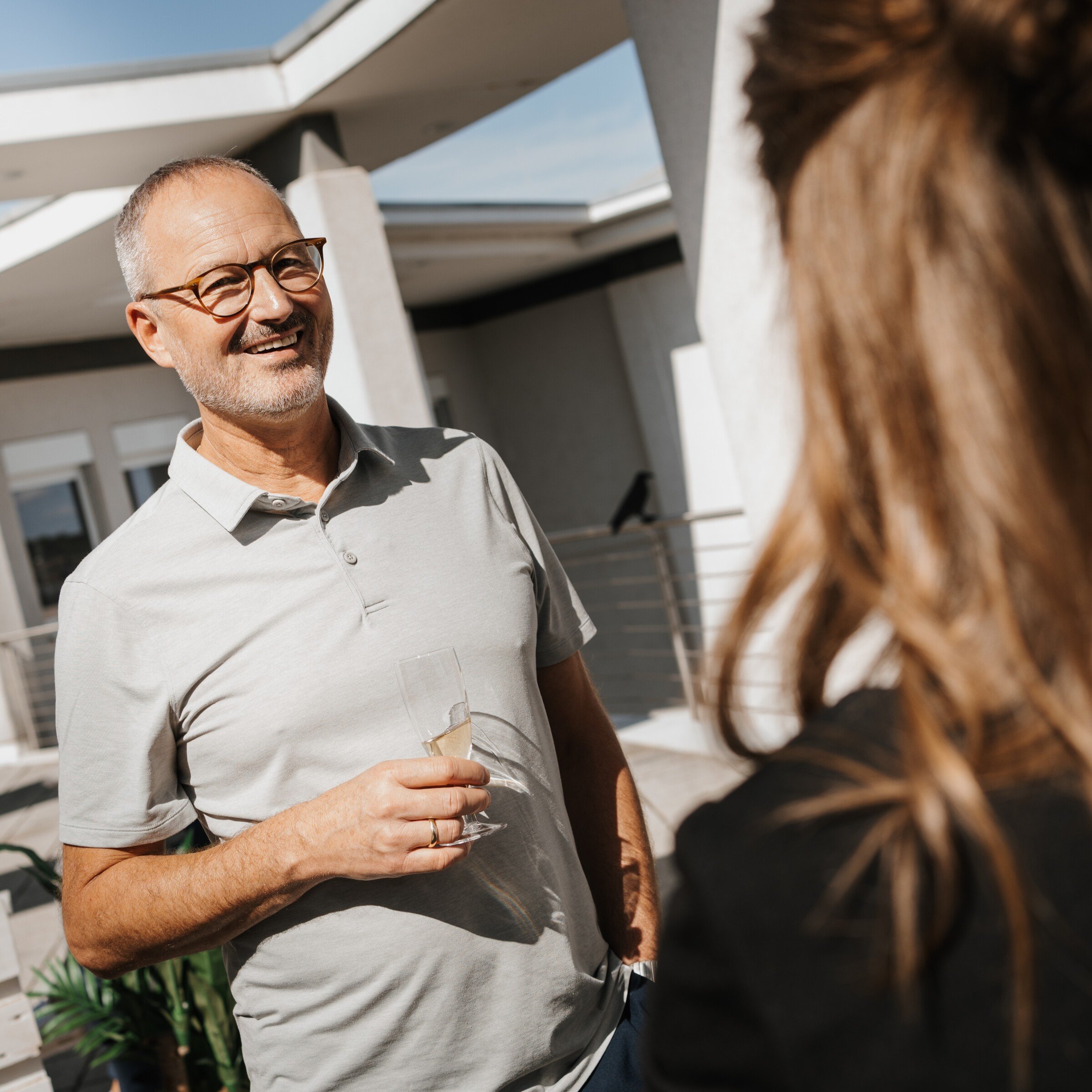 Foto eines Mannes mit grauem Hemd, Getränk in der Hand, der eine Frau gegenüber anlächelt.